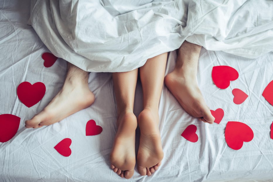 Fotografija: Cropped image of young couple is lying on bed. Close up of male and female feet. Loving couple is lying on bed under blanket covered by small red paper hearts. Saint Valentines Day.