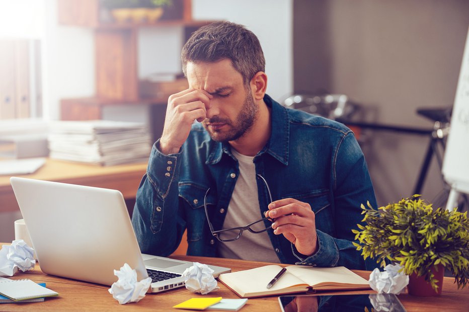 Fotografija: Frustrated young man massaging his nose and keeping eyes closed while sitting at his working place in office FOTO: G-stockstudio Getty Images/istockphoto