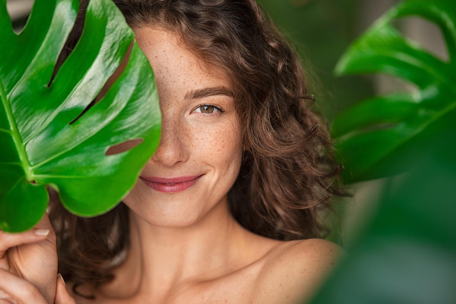 Fotografija: Close up face of beautiful young woman covering her face by green monstera leaf while looking at camera. Natural smiling girl with green palm leaf. Portrait of beauty woman with natural makeup and freckles on skin standing behind big green leaves.