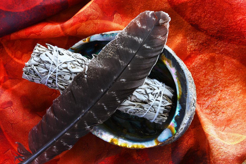 Fotografija: An image of an abalone shell with white sage bundle and sacred feather on a bright red background.