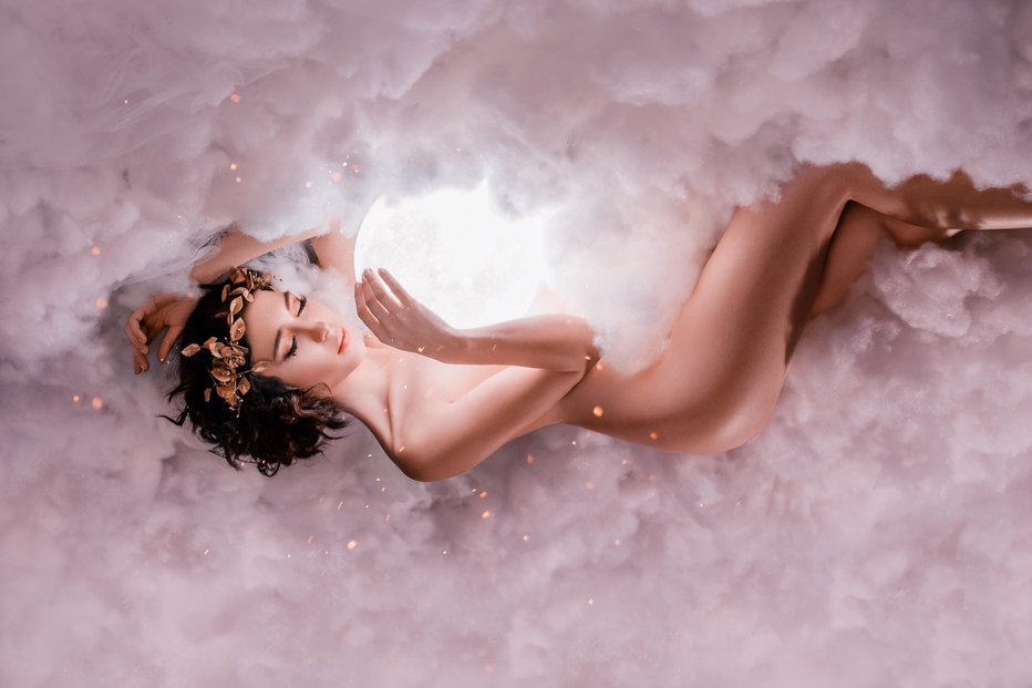 Fotografija: the night and day goddess sleeps in the clouds, as in a thick white fog, hugs the full moon, a naked nude sexy girl with a golden wreath and makeup, with childish innocent face in the glare of fire.
