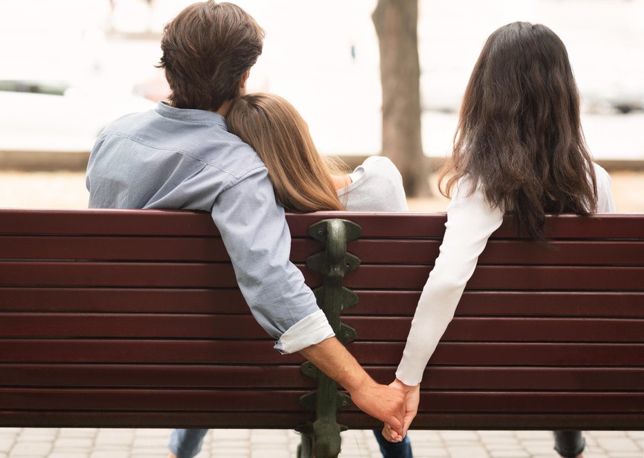 Fotografija: Love Triangle. Cheating Boyfriend Hugging Girlfriend Holding Hands With Her Girl Friend Sitting On Bench Together In Park Outdoor. Back-View
