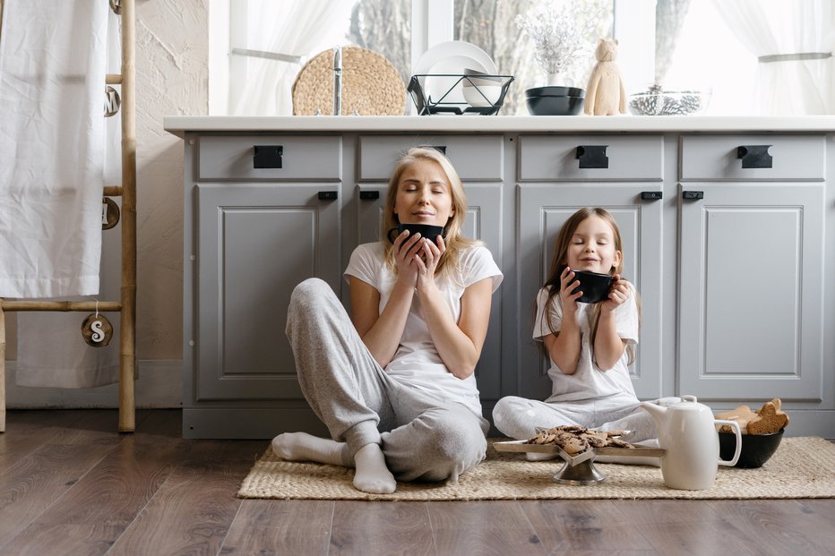 Fotografija: Dreamy mother sitting on kitchen floor together with her lovely daughter, enjoying morning coffee, drinking beverage and spending weekend in cozy apartment, resting at home