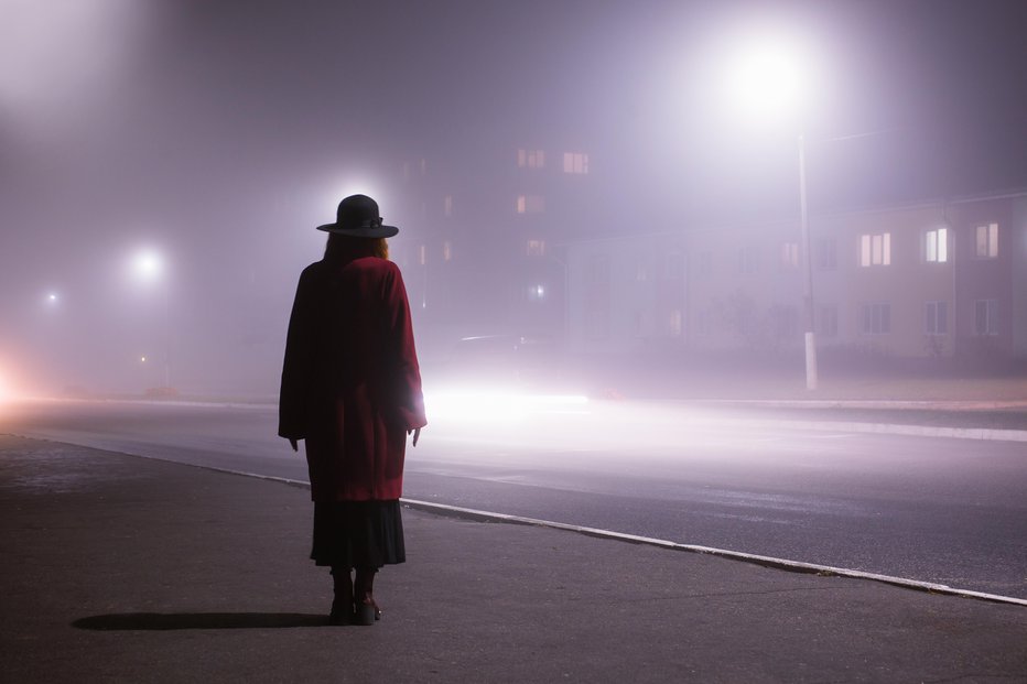 Fotografija: Woman silhouette on background of the night city in fog. Thick mist in dark scary evening city. Dark noir silhouette in hat on background of fog. Alone woman in mist. Noir mystery film. Lost man