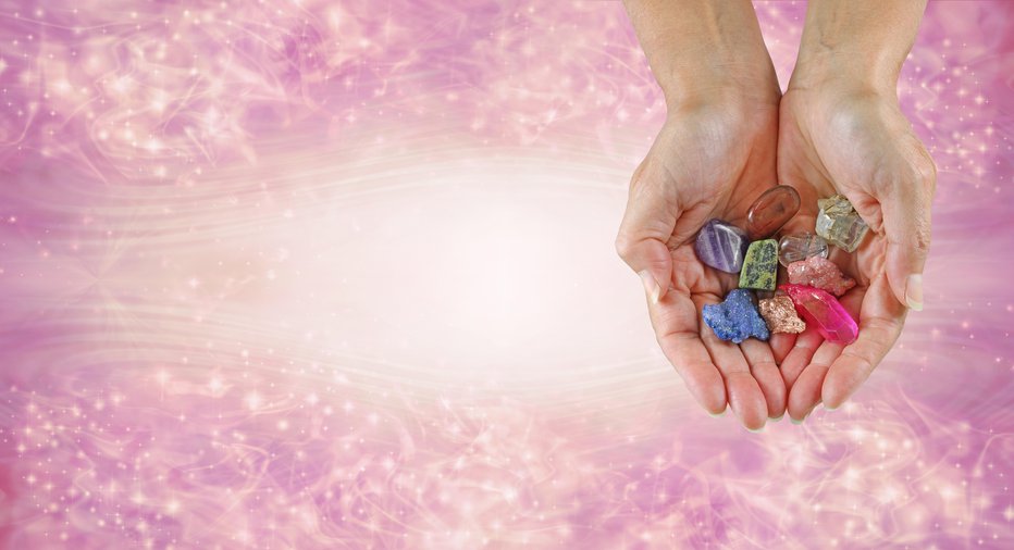 Fotografija: female hands cupped with a selection of raw and tumbled healing stones against a pink sparkling flowing background with space for text